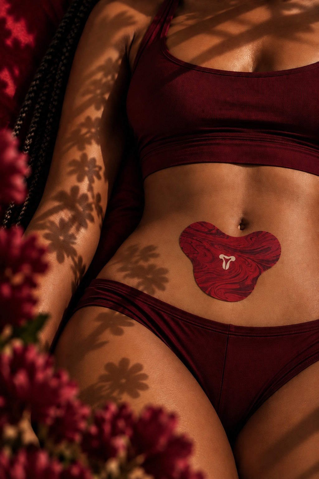 A woman lying in warm low light with the marbled period patch on her lower abdomen, flower shadows falling across her skin